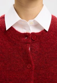 Cárdigan rojo de punto con una textura suave, que presenta un escote redondeado, dos botones rojos y una camisa blanca con cuello en capas debajo.