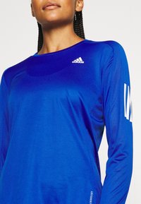 Sininen pitkähihaiseen urheiluprintti, joka on valmistettu hengittävästä kankaasta. Paidassa on Adidas-logo rinnassa ja tyyliteltyjä valkoisia yksityiskohtia hihoissa.