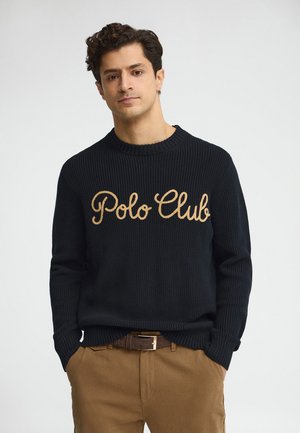 Uomo con capelli scuri e ricci che indossa un maglione nero lavorato a maglia con la scritta "Polo Club" e pantaloni marroni, con le mani in tasca.