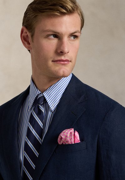 Tengerészkék zakó, kék-fehér csíkos ing, átlós csíkos nyakkendő és rózsaszín mintás zsebkendő. Sima szövet textúra és szabott fazon.