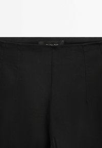 Pantalon noir ajusté avec des plis marqués, une ceinture sans coutures et une petite étiquette de marque visible à l'arrière.