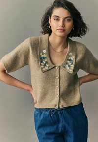 Kortärmad beige kofta med en bred krage, prydd med blommig broderi i blått och vitt, samt framknäppningar. Matcher med blå byxor.