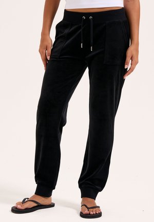 Schwarze Velours-Jogginghosen mit elastischem Bund, seitlichen Taschen, schmal zulaufenden Bündchen und einer weichen Textur. Kombiniert mit schwarzen Flip-Flops.