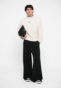 Helle beige Kapuzenpullover mit kleinem Logo, kombiniert mit schwarzen weiten Hosen und schwarzer Tasche. Verfügt über einen übergroßen Schnitt und strukturierten Stoff.
