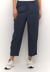 Pantalon court bleu marine avec plis à l'avant, en tissu léger, associé à des baskets blanches et grises.
