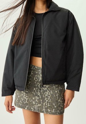 Vrouw draagt een zwarte jas met rits over een zwart crop top, gecombineerd met een groene en grijze minirok met luipaardprint.