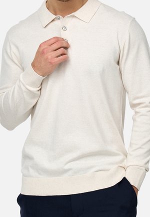 Homme portant un polo crème à manches longues, boutonnant le bouton supérieur avec la main gauche, la main droite dans la poche d'un pantalon marine.