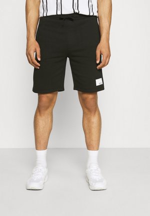 Mężczyzna ubrany w czarne szorty ze ściągaczem i białym prostokątnym logo na lewej nogawce, białe skarpetki, białe trampki oraz białą koszulę z pionowymi czarnymi paskami.