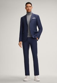 Marineblauer Maßanzug mit einem grauen Rollkragenpullover, verziert mit einem weißen Taschentuch. Schlanker Schnitt, glatte Textur, kombiniert mit weißen Sneakern.