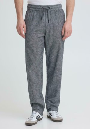 Uomo con pantaloni grigi con coulisse e sneakers bianche con strisce nere, in piedi contro uno sfondo chiaro uniforme.