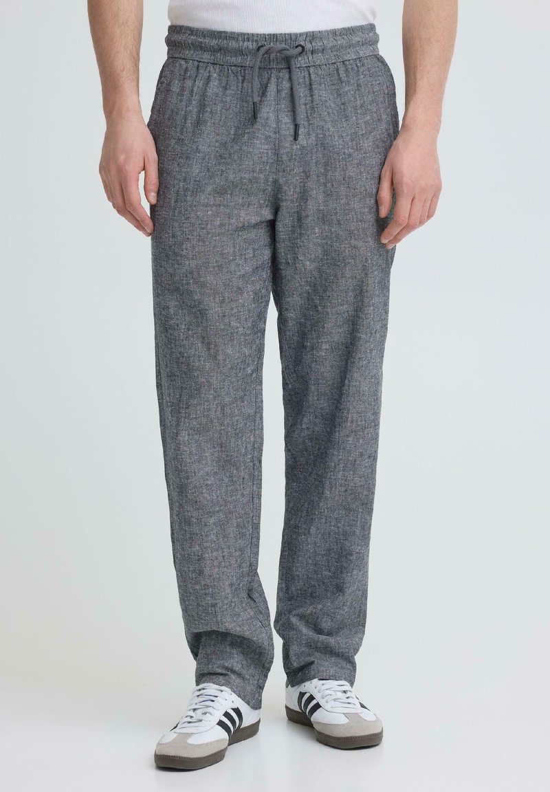 Άνδρας που φοράει γκρι παντελόνι με κορδόνι και λευκά αθλητικά παπούτσια με μαύρες ρίγες, στέκεται μπροστά σε ένα απλό φωτεινό φόντο.