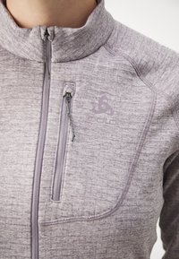 Lys lilla zip-up jakke laget av teksturert stoff, med høy krage, frontlomme med glidelås og subtilt logo på brystet.