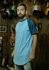 Světle modré oversized tričko s tmavě modrými raglánovými rukávy, zaobleným lemem, hladkým materiálem, nošeno s světle modrými džíny. V pozadí jsou helmice a dekorace.