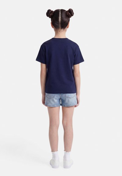 Παιδί με κότσιαδικο χτένισμα, φορώντας μπλε μπλούζα, ανοιχτόχρωμο τζιν σορτς, λευκές κάλτσες και αθλητικά παπούτσια, στέκεται γυρισμένο με την πλάτη σε λευκό φόντο.