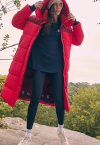 Raudona pūkinė striukė vidutinio ilgio, su gobtuvu ir kontrastinga vidine pamušalu. Dėvima virš juodos bluzonės ir juodų timpos, derinama su sportbačiais.