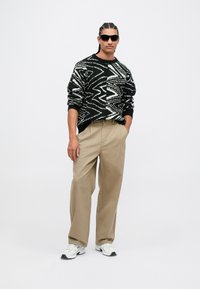 Mann trägt einen schwarz-weißen Zickzack-Pullover, beigen weiten Hosen, weiße Sneakers und schwarze Sonnenbrille und steht mit den Händen in den Taschen.