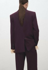 Blazer burdeos con diseño dividido, con mangas largas, puños abotonados y una textura suave, acompañado de pantalones a juego.