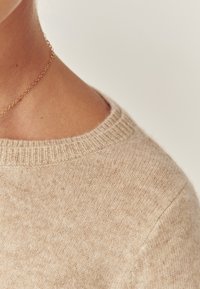 Beige gebreide trui met een ronde halslijn, met een zachte textuur en subtiele vlekjes verspreid over de stof. Gouden ketting zichtbaar bij de hals.