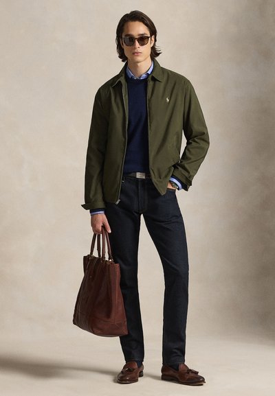 Olivno zelena jakna, mornarsko modra majica, modra s ovratnikom, temne kavbojke, rjava usnjena torba in rjavi čevlji. Model pozira pred nevtralnim ozadjem.