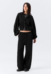 Γυναίκα με φαρδιά μαύρη παντελόνα, cropped μαύρο hoodie με φερμουάρ πάνω από ριγέ πουκάμισο, στέκεται με τα χέρια στις τσέπες, κοιτάζοντας προς τα πλάγια.