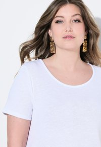 Bílá tričko s krátkými rukávy a kulatým výstřihem. Modelka má na sobě velké zlaté strukturované náušnice. Vlasy má upravené do jemných vln.