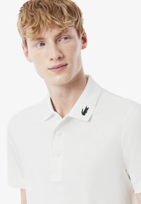 T-shirt polo blanc avec un col texturé, doté d'un logo de crocodile vert sur le côté gauche, et de trois boutons sur le devant.