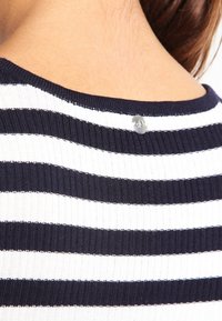 Gestreepte trui met een geribbelde structuur, marineblauwe en witte strepen, en een metalen logoaccent op de achterkant van de halslijn.
