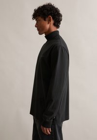 Μαύρο μπλουζάκι με μακριά μανίκια και ψηλό γιακά, μαλακή υφή, μινιμαλιστικός σχεδιασμός και διακριτικό λογότυπο στο στήθος. Συνδυάζεται με σκούρα τζιν.