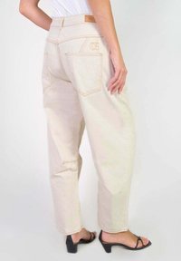 Jeans beiges taille haute avec une coupe décontractée, présentant des coutures contrastées, deux poches arrière et une étiquette en cuir brun issu de marque sur la ceinture.