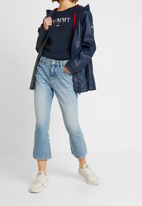 Laivastonsininen vedenpitävä takki punaisilla yksityiskohdilla, yhdistettynä laivastonsiniseen T-paitaan ja vaaleansinisiin leveälahkeisiin farkkuihin; valkoiset lenkkarit täydentävät asun.