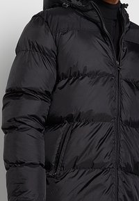 Veste matelassée noire avec poches zippées portée sur un pull à col roulé noir, partiellement zippée à l'avant.