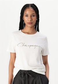 Bílý bavlněný tričko s lesklým stříbrným designem textu "Champagne". Krátké rukávy a klasický kulatý výstřih.