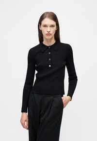 Γυναίκα που στέκεται με το χέρι στην τσέπη, φορώντας μαύρο μακρυμάνικο πλεκτό πόλο με κουμπιά και μαύρο πιετεντέ παντελόνι, μπροστά σε λευκό φόντο.