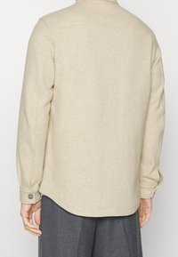 Beige wollenshirtjack met een afgeronde zoom, zachte textuur, lange mouwen en een knoopsluiting bij de manchetten, gezien van achteren.