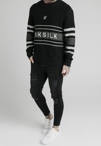 Svart stickad tröja med vitt randigt mönster och logotyp på framsidan, kombinerad med slitna svarta jeans och vita sneakers med guldaccenter.