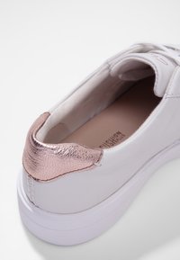 Sneaker blanche avec une tige en cuir lisse, accent de talon texturé en rose gold, et une semelle en caoutchouc blanche. L'intérieur présente un détail de rembourrage.