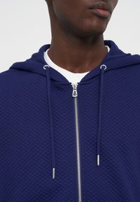 Άνδρας που φοράει μπλε σκούρο φερμουάρ με επένδυση και κορδόνια πάνω από λευκό μπλουζάκι με στρογγυλή λαιμόκοψη, εμφανιζόμενος από τους ώμους μέχρι το πηγούνι.