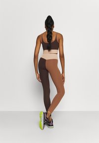 Brown and beige activewear set featuring a fitted sports bra and leggings with color-blocking design. Model wears purple shoes with green soles.