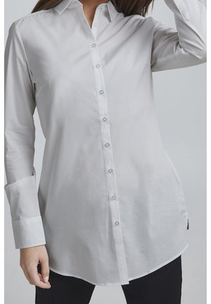 Kobieta ubrana w długą białą koszulę na guziki z długimi rękawami, stojąca na tle jednolitego jasnego tła.