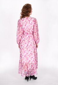 Lang, vloeiende jurk van doorschijnende stof met een roze bloemenpatroon. Heeft lange mouwen, een strikceintuur in de taille en een bescheiden halslijn.