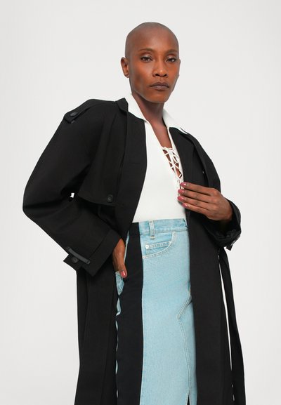Cappotto lungo nero con maniche ampie, chiuso con bottoni. Sotto, una maglietta bianca con scollatura in pizzo e una gonna di jeans azzurro chiaro.