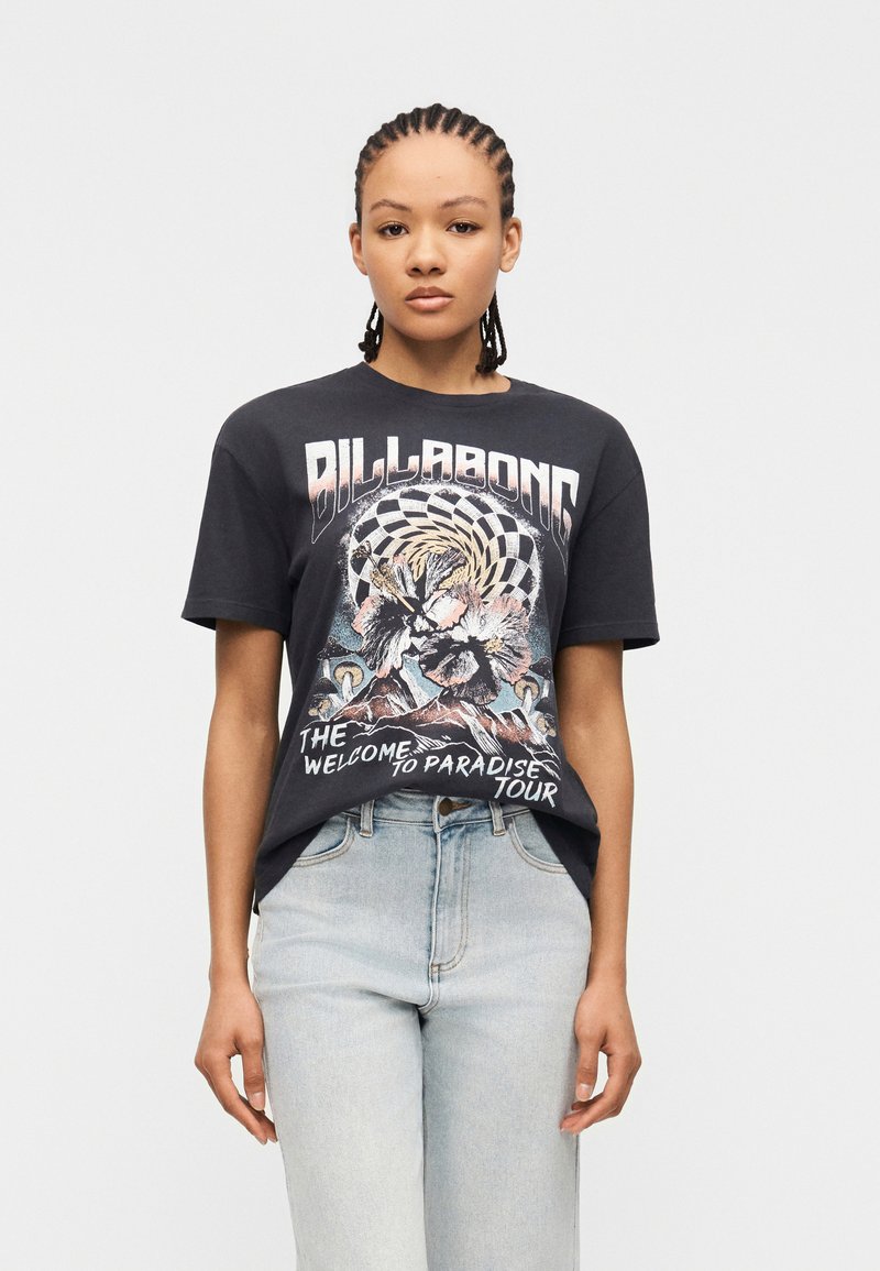 Νεαρό άτομο με πλεξούδες, φοράει μαύρο μπλουζάκι Billabong με γραφικά και ανοιχτό μπλε τζιν ψηλόμεσο, σε απλό φόντο.