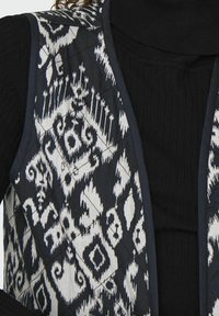 Quilted prsluk u tamnoplavoj boji s bijelim ikat uzorkom, sa glatkom teksturom i slojevitim dizajnom, nosi se preko crne rebraste majice s visokim ovratnikom.