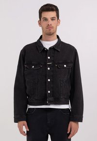 Sort denimjakke med en cropped design, knaplukning, to brystlommer og kontrasterende syninger, båret over en hvid skjorte.