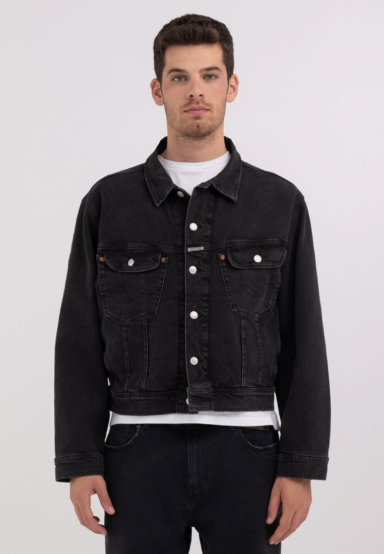 Sort denimjakke med en cropped design, knaplukning, to brystlommer og kontrasterende syninger, båret over en hvid skjorte.