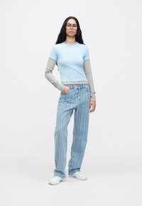 Γυναίκα με μακριά σκούρα μαλλιά και γυαλιά, που φοράει στρωματοποιημένα μπλε και γκρι τοπ, χαλαρούς ριγέ τζιν και λευκά αθλητικά παπούτσια, στέκεται με το ένα χέρι στην τσέπη.