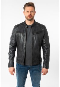 Schwarze Leder-Bikerjacke mit gesteppten Schulterakzenten, zwei Reißverschluss-Brusttaschen und einem Stehkragen. Kombiniert mit einem grauen T-Shirt und Jeans.