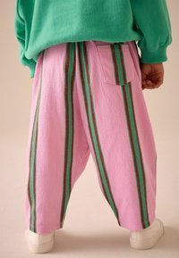 Heledad roosad püksid, millel on vertikaalsed rohelised ja punased triibud; tekstuuriga kangas; mugav lõige; tagatasku nähtav; kombineeritud valgete kingadega.