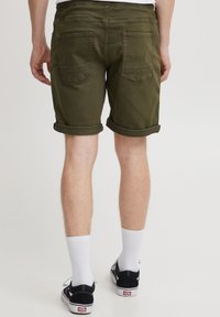 Olíva zöld pamut rövidnadrág felhajtott szegéllyel, két hátsó zsebbel és sima textúrával, fekete tornacipővel és fehér zoknival viselve.