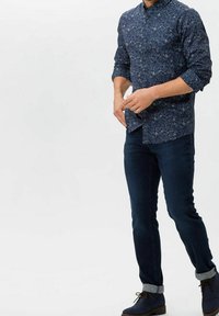 Man wearing dark blue floral button-up shirt, dark rolled-up jeans, and navy lace-up boots, standing against white background.